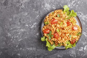 Fusilli pasta with tomato sauce, cherry tomatoes, lettuce and herbs on a black concrete background.