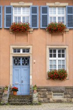 Picture of a traditional house with blue front door and shutters, decorated with red geraniums in