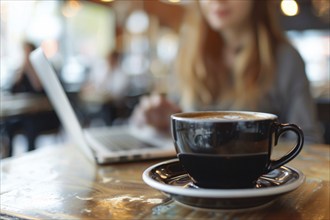 Black cup of coffee on table next to woman working on laptop in restaurant. Generative Ai, AI