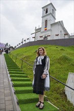 Young woman in folk traditional costume in front of a church on a green hill, woman in traditional