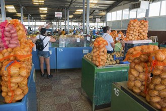 A market stall with potatoes in net bags, customers shopping in the background, market hall,