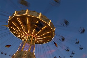 An illuminated carousel at dusk, spinning fast, in front of a blue sky with clouds and a Germany