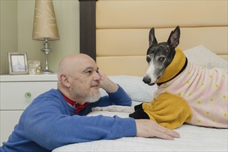 A mature man gazes lovingly at his pet, a Greyhound wearing an orange coat to keep warm, during a