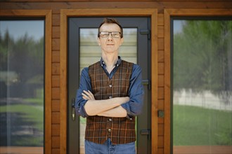 Middle-aged man in blue shirt and brown vest stands confidently in front of modern wooden house