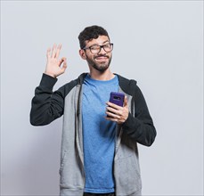 Pleasant person holding cellphone and winking, smiling boy with cellphone making ok sign, concept