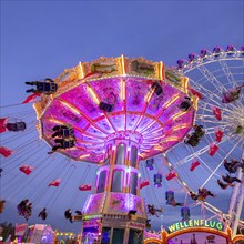 A lively funfair at dusk, illuminated by a large Ferris wheel and a colourful chain carousel,