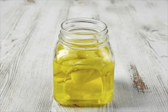 A square glass jar on a white wooden table. The jar is filled with a pickled yellow Chilli peppers