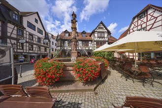 Market fountain with geraniums and outdoor area of a restaurant under a blue sky with cumulus