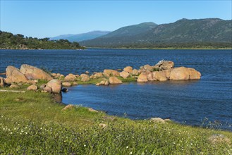 Landscape with calm lake surrounded by rocks and meadows, mountains in the background under a clear