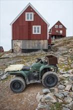 A green quad bike or ATV stands in front of red, wood-clad houses on a rocky hill, remote Arctic