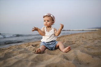 A thoughtful baby in summery clothes and flowers in her hair sits on the beach at sunset