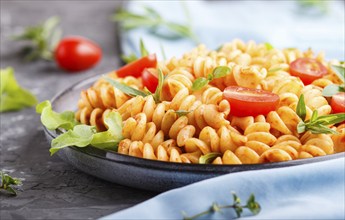 Fusilli pasta with tomato sauce, cherry tomatoes, lettuce and herbs on a black concrete background.