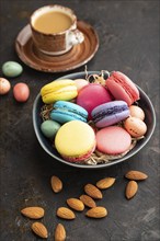 Multicolored macaroons and chocolate eggs in ceramic bowl, cup of coffee on black concrete