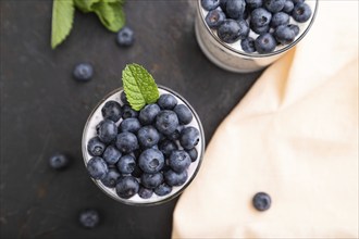 Yogurt with blueberry and chia in glass on black concrete background and orange linen textile. top