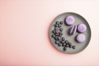 Purple macarons or macaroons cakes with blueberries on ceramic plate on a pastel pink background.