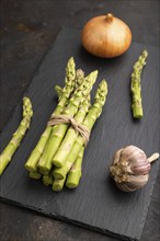 Bunch of fresh green asparagus, garlic, onion on slate board on black concrete background. Side