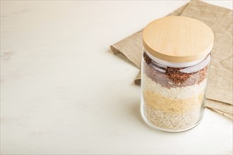 Glass jar with different kinds of rice poured in layers on white background. side view, copy space.