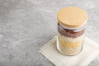 Glass jar with different kinds of rice poured in layers on a gray concrete background. side view,