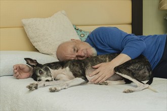 A bald man with a white beard enjoys a nap with his greyhound dog, lying in bed