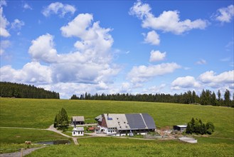 Farmstead with solar cells in a landscape with meadows and forest under a blue sky with cumulus