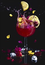 A colourful splashing cocktail with lemon slices, surrounded by flowers, reflection, dark