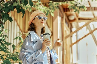 Stylish woman in blue raincoat and hat with a glass of coffee in hand looks at town district