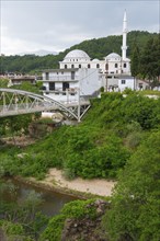 A mosque with a white dome and minaret next to a bridge over a river, surrounded by greenery,