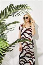 Beautiful woman in striped summer dress standing next to white wall surrounded by palm leaves