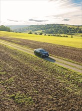 Black car driving on a gravelled country lane through a wide, rural landscape under a cloudy sky,