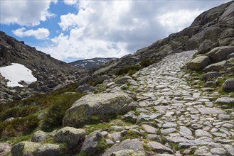 A stony hiking trail leads through a rocky mountain landscape under a partly cloudy sky, hiking