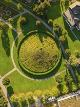 Aerial view of a circular Celtic burial mound in a green park, surrounded by trees and houses,