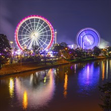 Two illuminated Ferris wheels are reflected in the water at night. The colourful scenery radiates