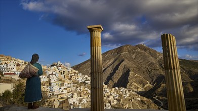 A life-size doll of a woman looks at the landscape framed by ancient columns, with a village in the