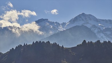 Snow-capped mountain peaks rise behind wooded hills under a blue sky, Erymanthos Mountains,