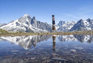 Young man doing a handstand in front of a mountain landscape, reflection in a small mountain lake,