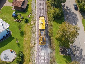 Yellow train runs along the railway tracks through the village next to a green meadow and houses,