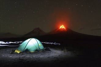 Night mountain landscape of Kamchatka Peninsula: illuminated tourist camping on background eruption