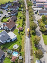 Aerial view of a train travelling through a residential area with green gardens and houses, tamping