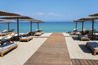 Wooden walkway leads to a sunlit beach with deckchairs and parasols by the clear sea, Lago Seaside