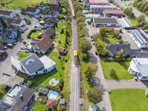 Yellow train runs through the village on the rails, surrounded by houses and green gardens, tamping