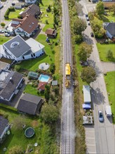 Yellow train runs on railway tracks through the village, surrounded by houses and gardens, tamping