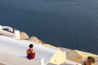 Unrecognised woman sitting on the white roof of a house looking at the sea. Santorini island in the