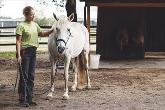 Caucasian woman trainer holding a white horse on the leash, taking hime horseback riding on the