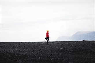 A traveler in a red jacket standing on a black sand beach in Iceland. One person only, cold foggy