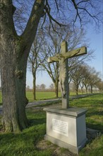 A stone crucifix next to a tree-lined path in a rural landscape under a blue sky, Borken, North