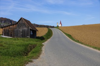 Autumn atmosphere, country road leads through freshly cultivated farmland, church tower and wayside