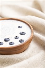 Yogurt with blueberry in wooden bowl on white wooden background and linen textile. Side view, close