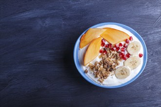 A blue plate with greek yogurt, granola, persimmon, banana, pomegranate black wooden background.