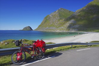 Bicycle with red bags on a road along a beach with turquoise water, in the middle of mountains and