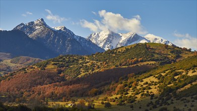 Autumn mountain landscape with snow-covered peaks under a cloudy blue sky, autumn foliage